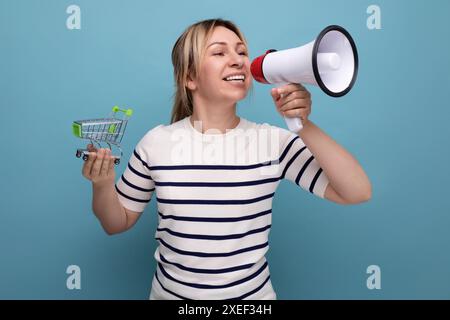 Blonde attraktive junge Frau in lässiger Kleidung spricht mit einem Megaphon, während sie einen Supermarkt-Trolley auf blauem Hintergrund hält Stockfoto