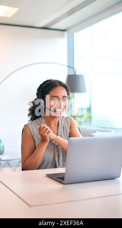 Frau Mit Headset, Die Am Schreibtisch Sitzt Und Mit Laptop Gute Nachrichten Feiert Oder Das Verkaufsziel Erreicht Stockfoto