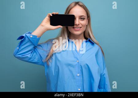 Lächelndes informelles blondes Mädchen, das Smartphone mit leerem Bildschirm für Website auf blauem Hintergrund hält Stockfoto