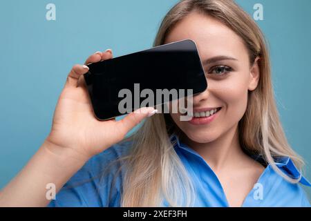 Lächelndes, stilvolles blondes Mädchen, das Smartphone mit Mockup für Webseite in der Nähe ihres Gesichts auf blauem Hintergrund hält Stockfoto