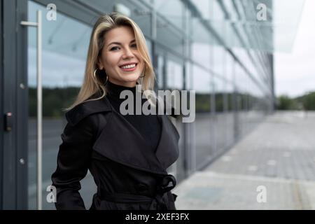 Glückliche junge Frau steht selbstbewusst vor dem Hintergrund eines modernen Gebäudes Stockfoto