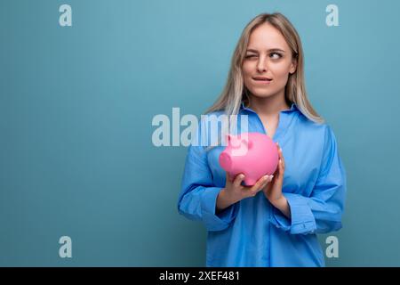 Kluges blondes Mädchen, das eine Schweinebank in Form eines Schweins mit Geld auf einem blauen isolierten Hintergrund hält Stockfoto