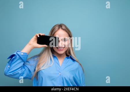 Lächelndes, attraktives blondes Mädchen, das leeres Smartphone-Display mit Modell für Webseite auf blauem Hintergrund zeigt Stockfoto