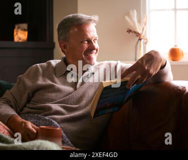 Reifer Mann Zu Hause Im Winter Pullover Auf Sofa Mit Warmem Heißem Getränk Kaffee Im Becher Lesebuch Stockfoto