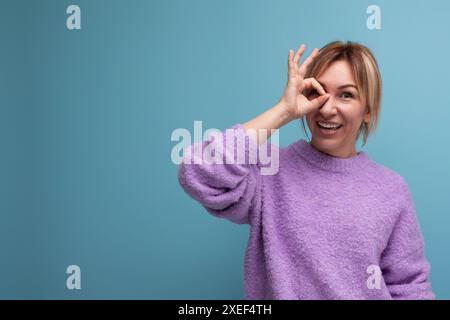 Nette charmante nette blonde junge Frau Beraterin in lila Pullover flirtet auf blauem Hintergrund mit Kopierraum Stockfoto