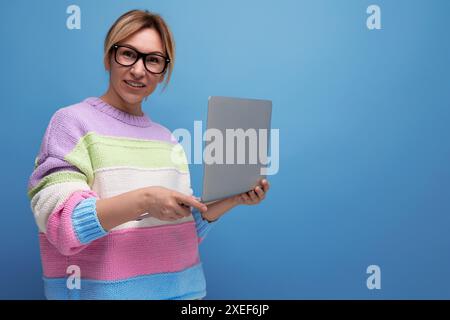 Die lächelnde blonde Beraterin in einer gestreiften Jacke arbeitet mit einem Laptop in den Händen auf einem blauen Hintergrund mit Kopierraum Stockfoto