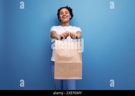 Junge glückliche Brünette mit Afro-Haaren in einem weißen Tanktop, die eine Bastelarbeit von einem Lieferdienst hält Stockfoto