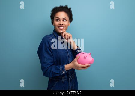 Junge fröhliche hispanische Geschäftsfrau mit flauschigem Haar in einem blauen Denim-Anzug hält Sparguthaben in einer Sparkasse Stockfoto