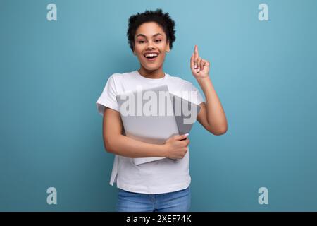 Junge glückliche Brünette mit Afro-Haaren in einem weißen Tanktop hält einen Laptop für die Arbeit in ihren Händen. Geschäftskonzept Stockfoto