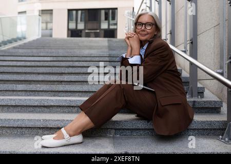 Geschäftsporträt einer selbstbewussten erfolgreichen 60-jährigen grauhaarigen Dame in Brille mit brauner Jacke Stockfoto