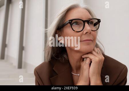 Das Porträt einer selbstbewussten, Reifen Dame mit grauen Haaren in Brille ist in einer braunen Jacke auf dem Hintergrund des Gebäudes gekleidet Stockfoto