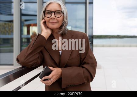 Selbstbewusste, gepflegte alte Dame mit grauem Haar in Brille in brauner Jacke wartet auf ein Treffen auf der Straße Stockfoto