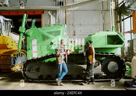 Extrem lange Aufnahme von ethnisch unterschiedlichen Männern und Frauen, die sich unterhalten, während sie eine Kaffeepause auf der Arbeit in modernen Schwermaschinenfabriken haben Stockfoto