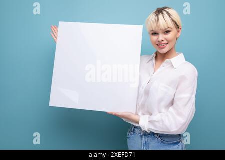 Junge erfolgreiche hübsche blonde Büroangestellte in weißem Hemd und Jeans hält ein Stück Papier, um eine Werbung einzufügen. Stockfoto
