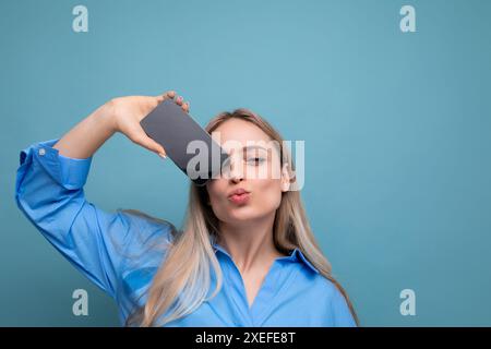 Lächelndes, attraktives blondes Mädchen, das Smartphone mit leerem Bildschirm für Webseite auf blauem Hintergrund hält Stockfoto