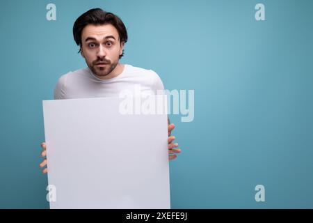 Junger brutaler brünette Mann zeigt Papierplakat mit Mockup Stockfoto