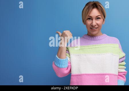 Blonde junge Frau in einem hellen Mantel zeigt mit dem Finger auf den Mauerraum auf blauem Hintergrund Stockfoto