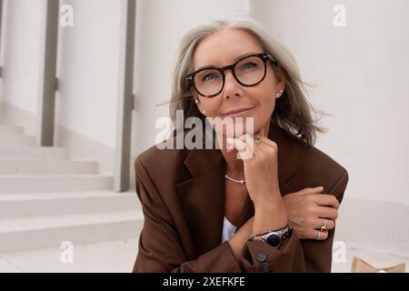 Eine schlanke Geschäftsfrau mit grauen Haaren und Brillen trägt eine Jacke, die auf der Treppe in der Nähe des Business Centers sitzt Stockfoto