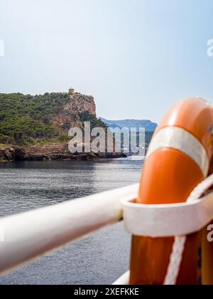 Rettungsschwimmer am Geländer eines Schiffes auf dem Weg nach Port de Sóller an einem ruhigen Frühlingstag mit dem Wachturm von Torre Picada in der Ferne. Stockfoto