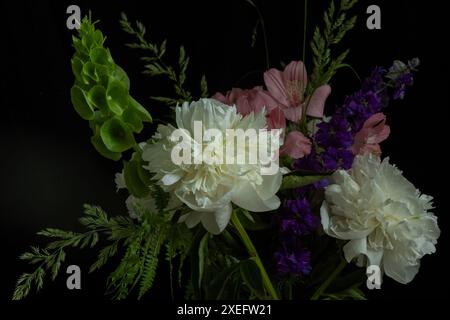 Eleganter Blumenstrauß mit weißen Pfingstrosen, rosa und lila Blüten und grünem Laub, der auf schwarzem Hintergrund für dramatische Momente angeordnet ist Stockfoto
