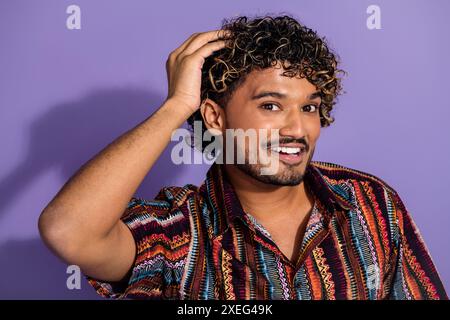 Foto von positivem nettem Mann mit Bart, der wellige Frisuren-gekleidetes Druckhemd im Friseurladen berührt, isoliert auf lila Hintergrund Stockfoto