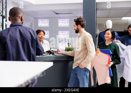 Mann in der Schlange, der kontaktlos per Smartphone bezahlt und beim Einkaufen am Black Friday mit elektronischer Kreditkarte bezahlt. Verschiedene Leute, die Kleidung halten, warten an der Kasse Stockfoto