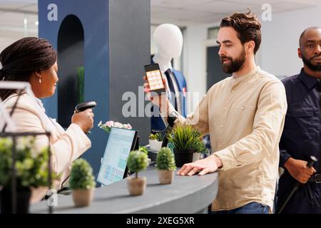 Mann Kunde mit Smartphone, der der Verkäuferin beim Einkaufen in einem Bekleidungsgeschäft einen Rabattcode anzeigt. Kassierer scannt Promo-Coupon, während Sie den Käufer am Checkout-Schalter servieren Stockfoto