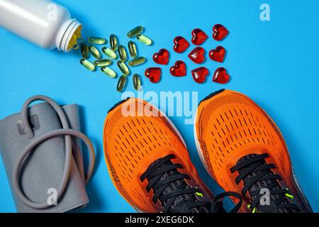 Tonometer zur Blutdruckmessung, sportliche orangefarbene Turnschuhe, gelbe Vitamin-D-Kapseln und rote Glasherzen auf blauem Hintergrund. Vitamin Stockfoto