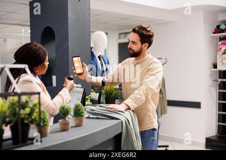 Kassierer aus afroamerikanischer Frau, die den mobilen Werbecode des Black Friday vom Smartphone des Kunden mithilfe eines Barcode-Scanners scannt. Junger Mann, der einen Gutschein einsetzt, um Rabatt beim Einkaufen in einem Bekleidungsgeschäft zu erhalten Stockfoto
