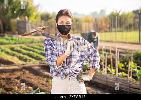 Asiatische Frau mit Schaufel im Gemüsegarten im Freien tragende Gesichtsmaske für Prävention Corona Virus Krankheit Stockfoto