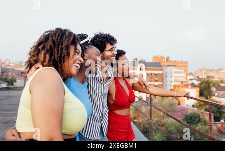 Vier fröhliche multiethnische Freunde schauen sich die Stadtlandschaft an. Süße afrikanische Frauen und Männer mit gemischter Rasse umarmen und Spaß auf dem Dach des Mult haben Stockfoto