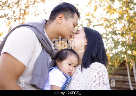 Ein verliebtes asiatisches Paar küsst sich draußen und hält sein Kind fest. Süße, liebevolle koreanische Eltern in lässiger Kleidung umarmen sie Stockfoto