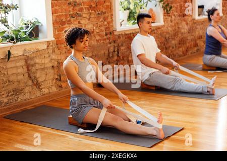 Eine Gruppe von sportlich attraktiven Menschen üben eine Yoga-Lektion mit einem Banderweiterer. Junge multiethnische Frauen und Männer sitzen Stockfoto