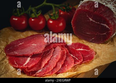 Ganze und geschnittene Bresaola auf Papier auf einem Schneidebrett mit Tomaten. Stockfoto