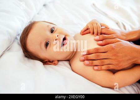 Ein süßes lächelndes Baby und seine Mom Hände. Eine Mutter berührt ihren kleinen Sohn in den Bauch. Ein lachendes süßes Baby liegt auf dem Bett. Stockfoto