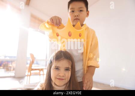 Niedliche Kinder verschiedener Rassen spielen zusammen. Ein kleiner asiatischer Junge hält eine Papierkrone über dem Kopf eines kaukasischen Mädchens. Stockfoto