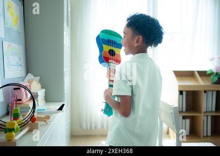 Kleiner Künstler, der die Ukulele im hinteren Teil des Klassenzimmers spielt, während er darauf wartet, dass Freunde das Klassenzimmer betreten Stockfoto