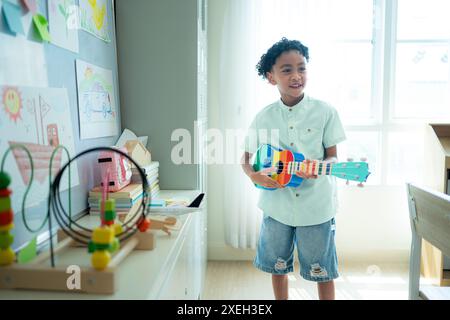 Kleiner Künstler, der die Ukulele im hinteren Teil des Klassenzimmers spielt, während er darauf wartet, dass Freunde das Klassenzimmer betreten Stockfoto