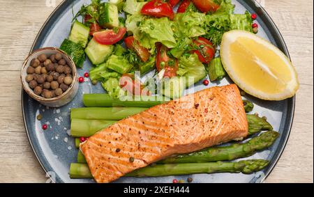 Gesundes Mittagessen mit gegrilltem Lachs mit Spargel und frischem Tomaten- und Gurkensalat, einer Reihe Zitronenscheibe. Essen auf einer blauen Runde Stockfoto