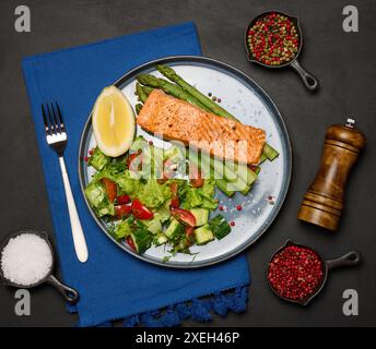 Gesundes Mittagessen mit gegrilltem Lachs mit Spargel und frischem Tomaten- und Gurkensalat, einer Zitronenscheibe. Essen auf einem blauen runden Teller Stockfoto