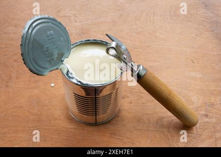 Geöffnete Metalldose mit Kondensmilch und Metalldosenöffner auf braunem Holzhintergrund. Camping-Food-Konzept. Stockfoto