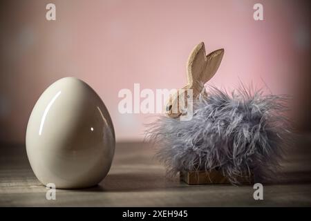 Eine dekorative Ostereinrichtung mit einem hölzernen Häschen mit Federn und einem Keramikei auf einer hölzernen Oberfläche mit rosa Hintergrund Stockfoto