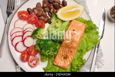 Gesundes Mittagessen mit gegrilltem Lachs auf grünem Salat, neben Gemüse, Tomaten, Radieschen, Brokkoli und einer Portion Bohnen. Stockfoto