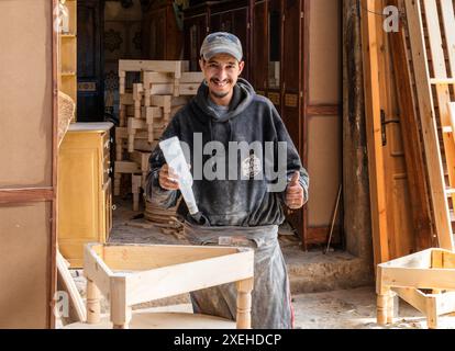 Ein junger Zimmermann lächelt stolz und arbeitet in seiner Holzwerkstatt und klebt einen Stuhl Stockfoto