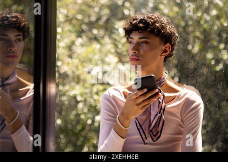 Smartphone in der Hand, junger Mann mit Schal, der zu Hause aus dem Fenster schaut Stockfoto