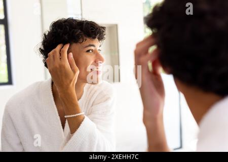 Ein lächelnder junger Mann im Bademantel, der auf das Spiegelbild im Badezimmerspiegel blickt Stockfoto