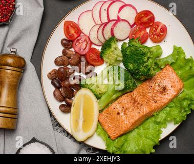 Gesundes Mittagessen mit gegrilltem Lachs auf grünem Salat, neben Gemüse, Tomaten, Radieschen, Brokkoli und einer Portion Bohnen. Stockfoto