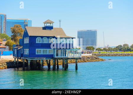 San Diego, Kalifornien - 16. April 2024: Blaues Holzhaus auf Stelzen über dem Wasser mit modernen Gebäuden von San Diego im Hintergrund. Stockfoto