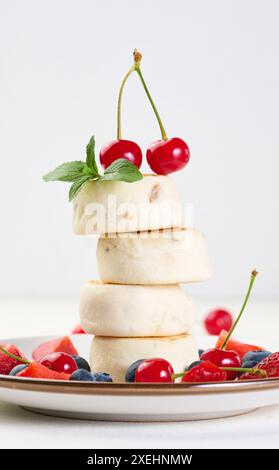 Ein Stapel von vier Käsekuchen, dekoriert mit zwei Kirschen und einem Minzzweig, auf einem Teller mit Beeren, Heidelbeeren, Erdbeeren Stockfoto