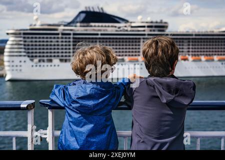 Zwei Jungs auf einem Fährdeck, die den Hafen von Kopenhagen verlassen. Ein Kind mit blonden Locken weist seinem Bruder etwas auf, während er ein Kreuzfahrtschiff an Land ansieht Stockfoto